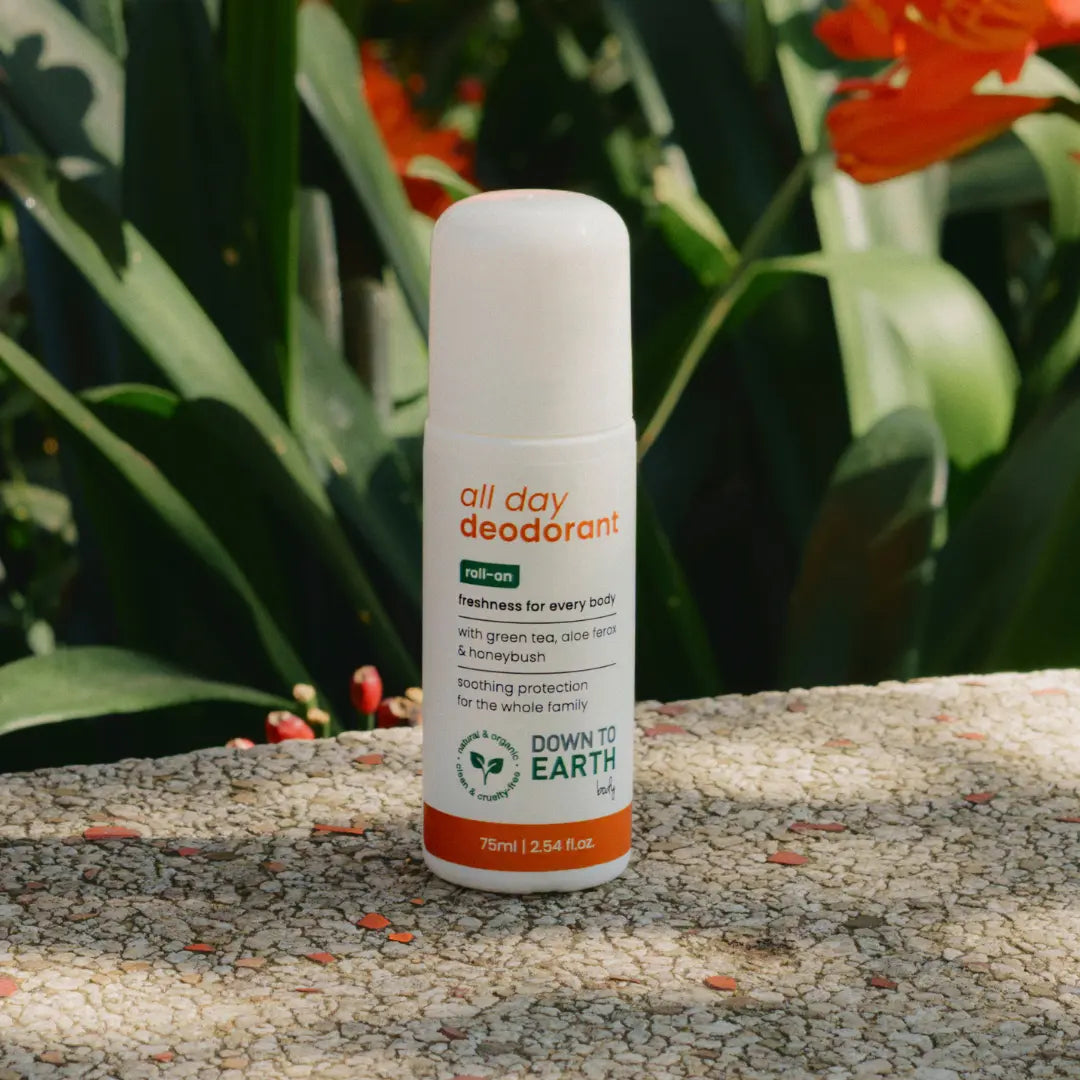 A 75ml roll-on deodorant bottle labeled 'all day deodorant' by Down to Earth, placed on a textured stone surface with lush green tropical leaves and orange flowers in the background.