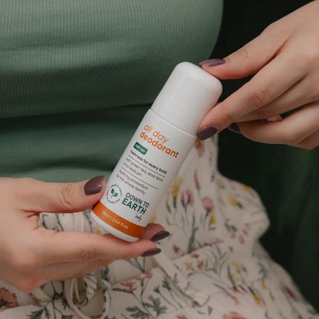 A person holding a white roll-on deodorant bottle labeled 'all day deodorant' and 'DOWN TO EARTH', with floral clothing visible in the background.
