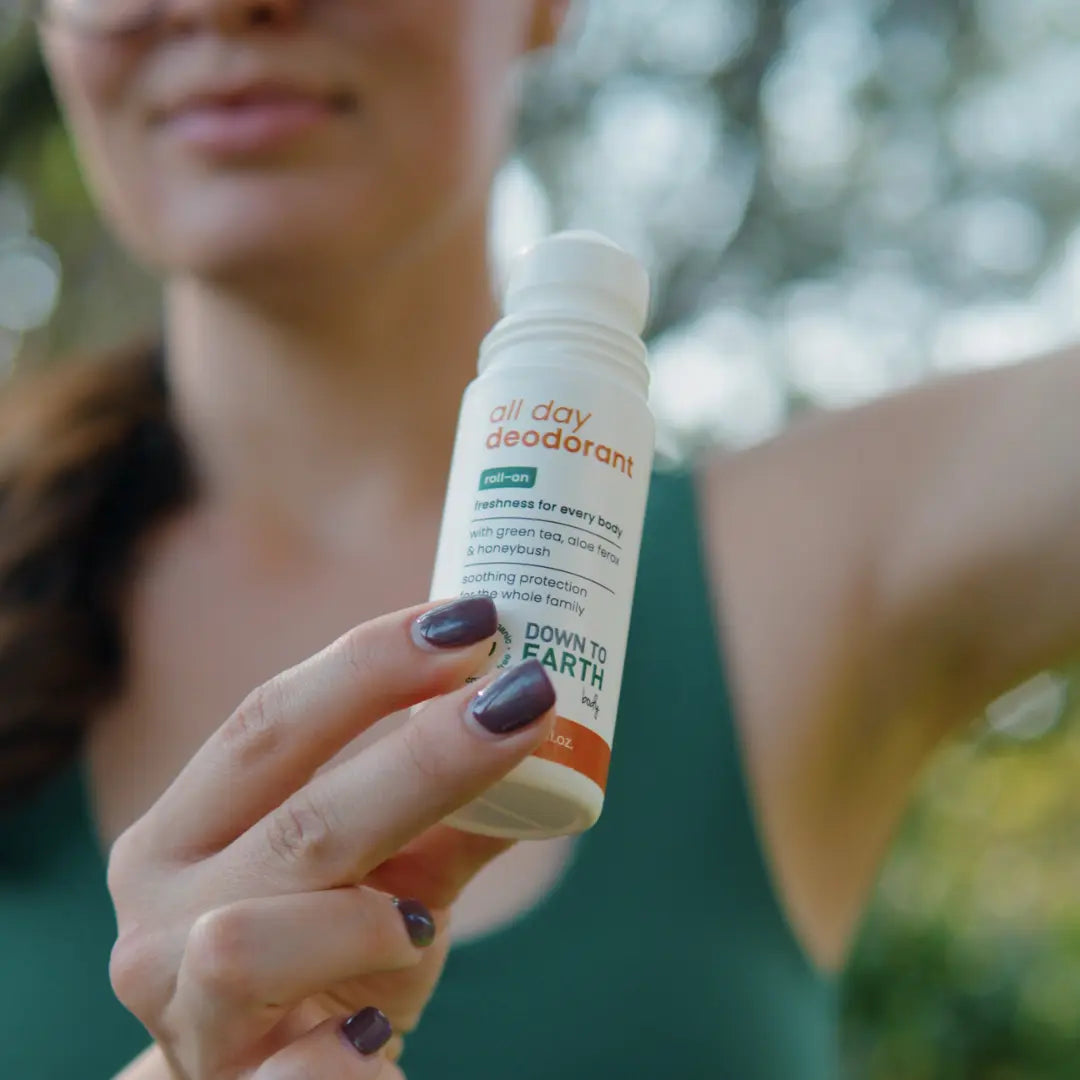 A person outdoors holds a small white roll-on bottle of deodorant. The label reads 'all day deodorant' and 'DOWN TO EARTH'.