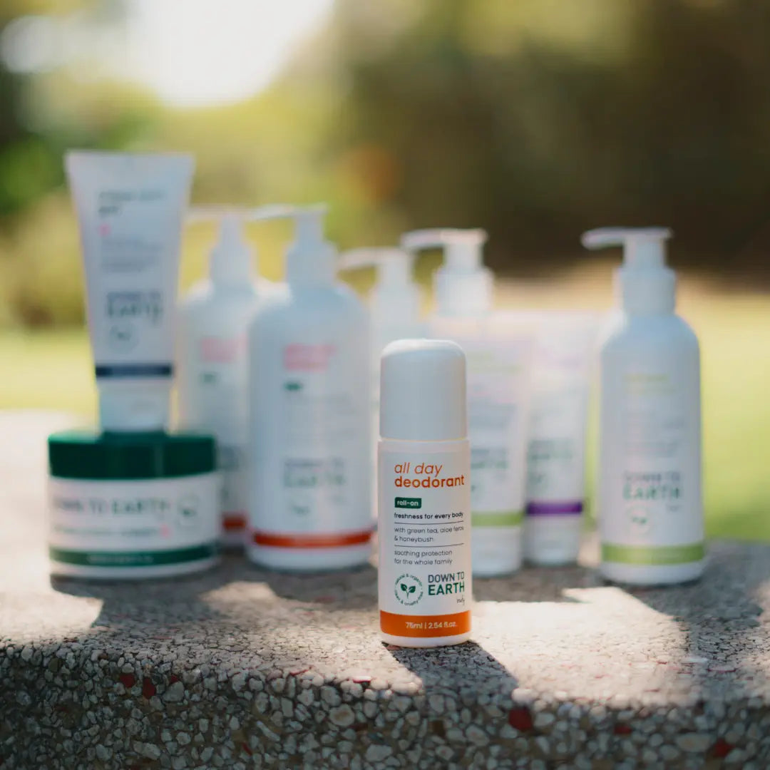 A bottle of Down to Earth All Day Deodorant stands in sharp focus in the foreground on a stone surface, with several other Down to Earth personal care products blurred in the background outdoors.