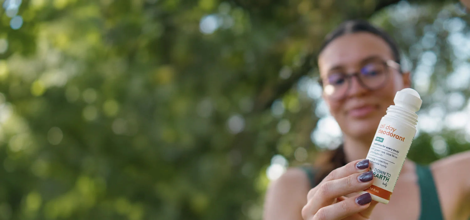 A woman outdoors holding a small white roll-on bottle with a white cap. The background is a soft, blurred green natural setting.