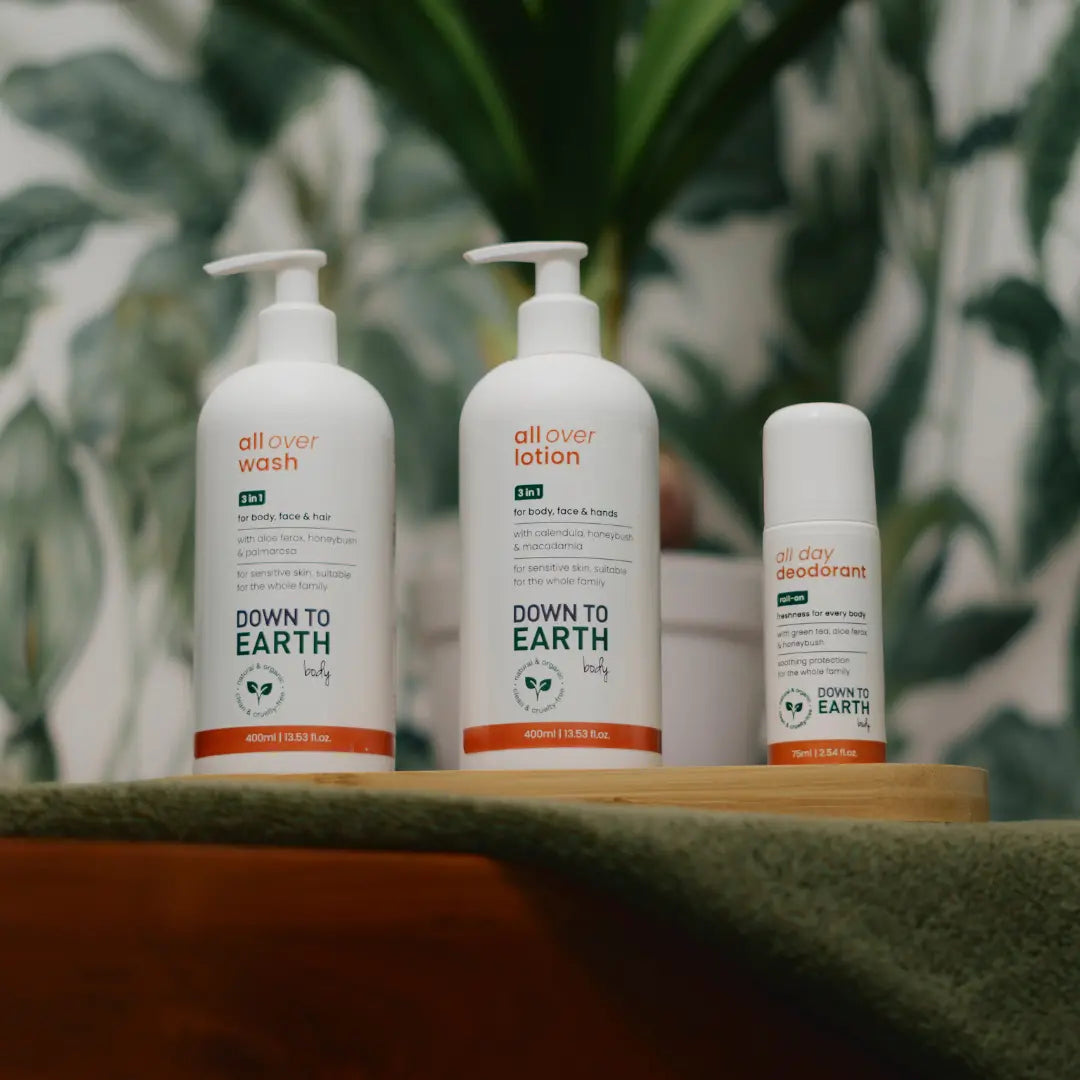 Three white skincare pump bottles and a white deodorant container from the brand 'all over' arranged on a wooden tray with a green textured cloth, set against a blurred tropical leaf background.