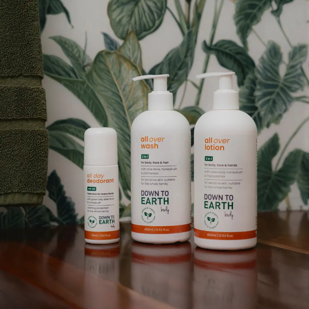 Three white pump bottles of Down to Earth Self-care products on a wooden surface against a tropical green leaf patterned wall. The bottles are labeled 'all over wash', 'all over lotion', and 'all clay deodorant'.