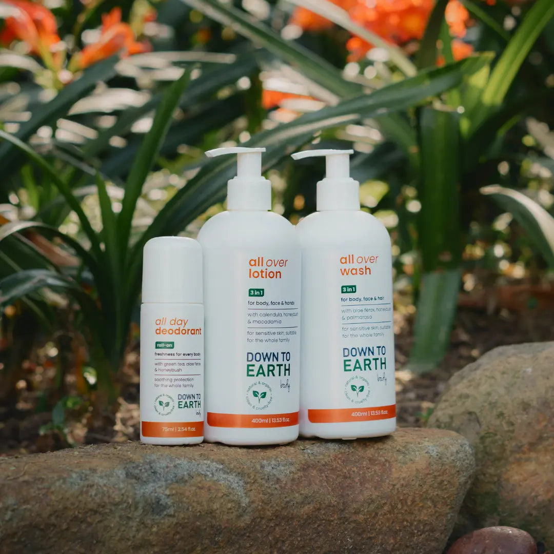 Three Down to Earth natural toiletry products on a stone surface with a blurred garden background of green leaves and orange flowers. The set includes a small deodorant bottle, a lotion bottle, and a wash bottle.