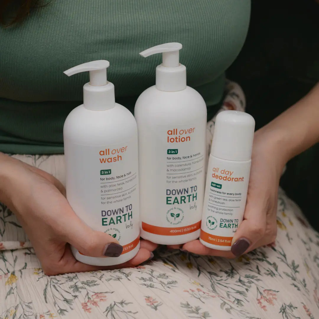 Three Down to Earth body care products held in hands: a large pump bottle of all over wash, a medium pump bottle of all over lotion, and a smaller roll-on deodorant, shown against a green top and floral fabric background.