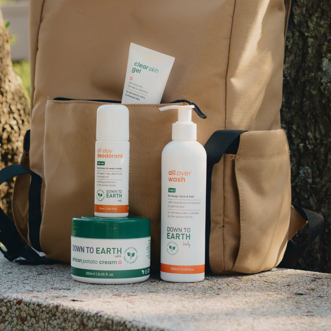 Three Down To Earth body care products arranged in front of a tan backpack outdoors on a stone surface: a white all day deodorant bottle, a white all over wash pump bottle, and a green-and-white steam potato cream tub.