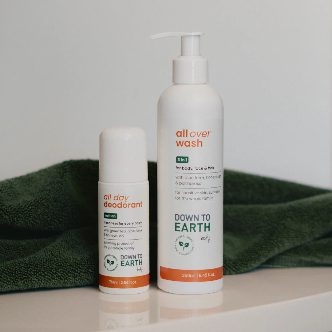 Two white Down To Earth body care products, an all day deodorant roll-on and an all over wash pump bottle, standing on a white surface with a folded dark green towel behind them.